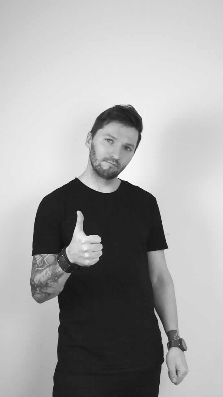 Grayscale footage of a white man with tattoos, short beard, hair and black clothing raising his arm and showing thumbs up expression with his thumb and fist showing that he likes something, is happy.