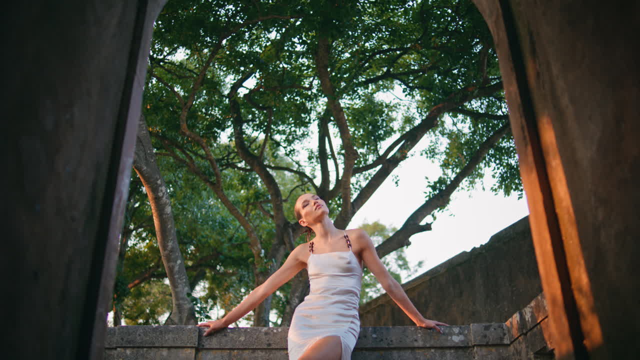 아치에서 돌 울타리에 기대어 고대 건물을 포즈하는 세련된 여인.