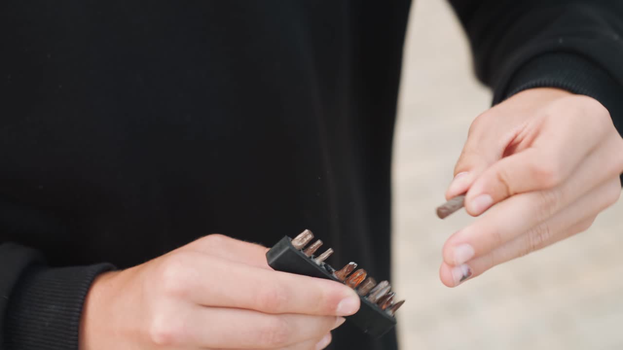 Tampilan dekat tangan memegang mata obeng oleh orang kulit putih di luar ruangan, teknisi jalanan memeriksa set logam, penempatan ujung jari yang fokus, pegangan taktil dan gerakan yang mantap, latar belakang beton di siang hari
