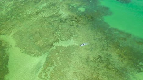 Drone-view-of-a-boat-sailing-in-the-natural-pools-formed-between-coral-reefs-on-Rua-Porto-Beach-in-Alagoas,-Brazil