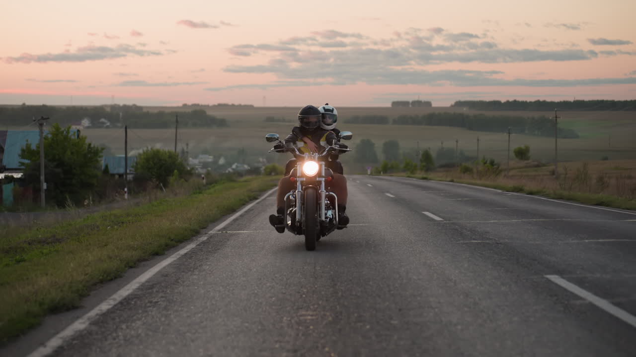Motorcyclist with passenger wearing helmets rides uphill on countryside road at sunset, headlight glowing against scenic horizon, rural fields and distant village visible under soft evening sky