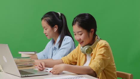 close up of asian woman students reading books and showing thumbs up gesture to the camera while sitting on a table with laptop in the green screen background classroom