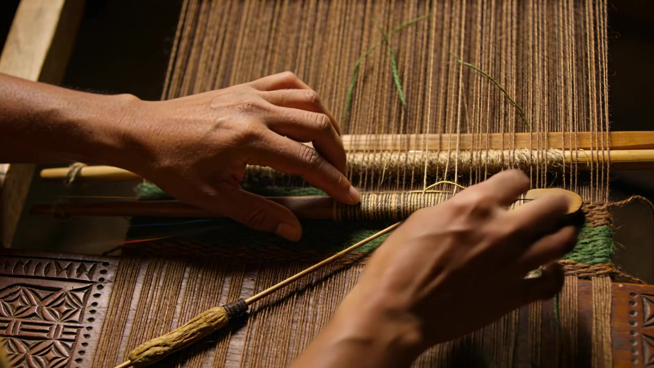 Entering hands, short-sleeved weaver guiding green shuttle through loom at bench making green strip