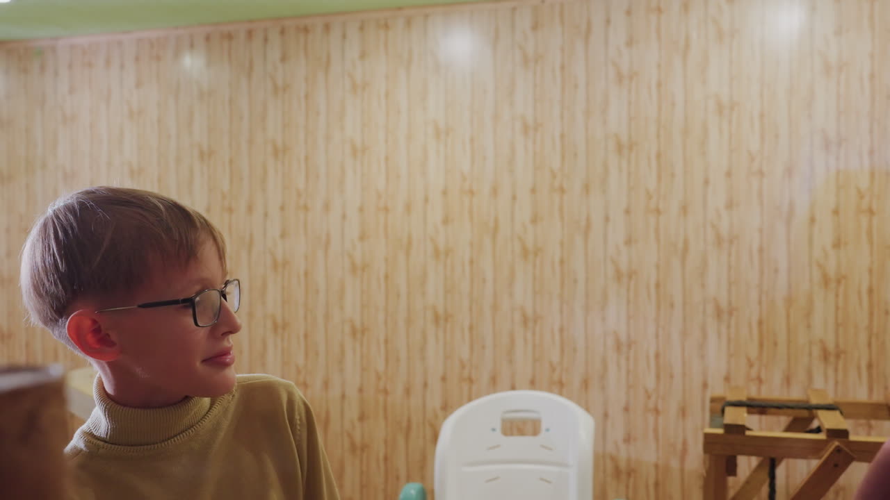 redhead woman caucasian boy sharing snack in cozy wooden kitchen, mother offers small treat to son in chair glasseswearing boy responds with grin while woman watches with gentle smile, warm lighting