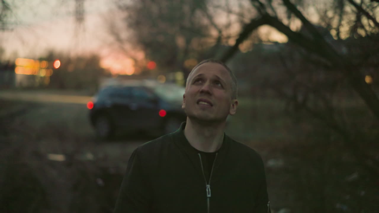A man in a green jacket stands in a dimly lit outdoor setting at dusk, looking upwards with a contemplative expression, smoke is visible around his nose and mouth