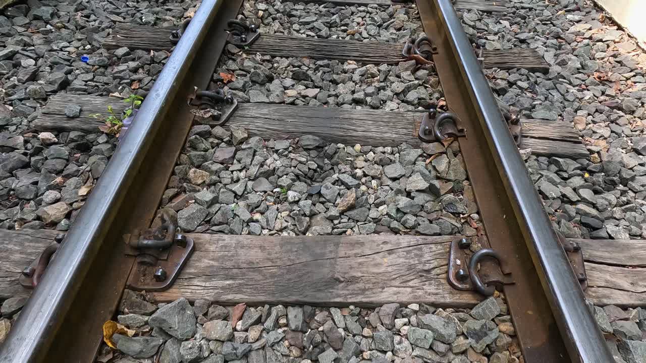 Point-of-view movement along metal railway tracks and wooden sleepers, surrounded by rocks, under natural daylight in Kanchanaburi, Thailand. Smooth forward camera motion