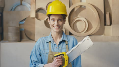 portrait of caucasian woman in helmet holding saw and smiling at camera in carpentry workshop