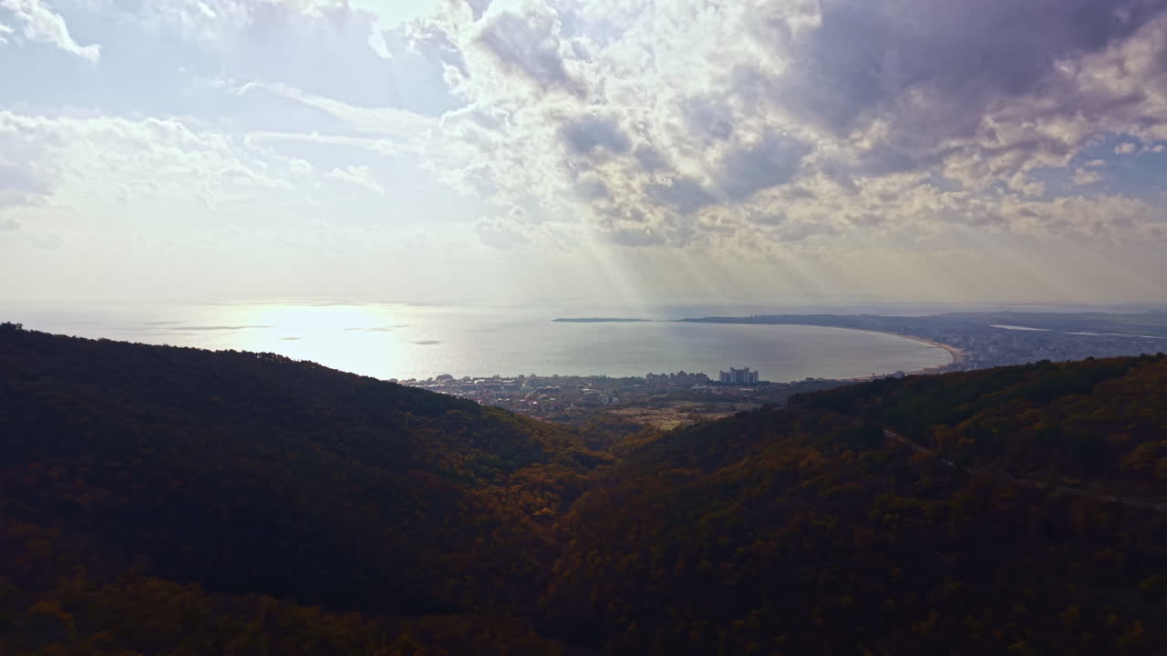 Vista panoramica sulla costa dalle montagne durante un sereno pomeriggio, con nuvole e sole che proiettano una luce soffusa sul mare scintillante