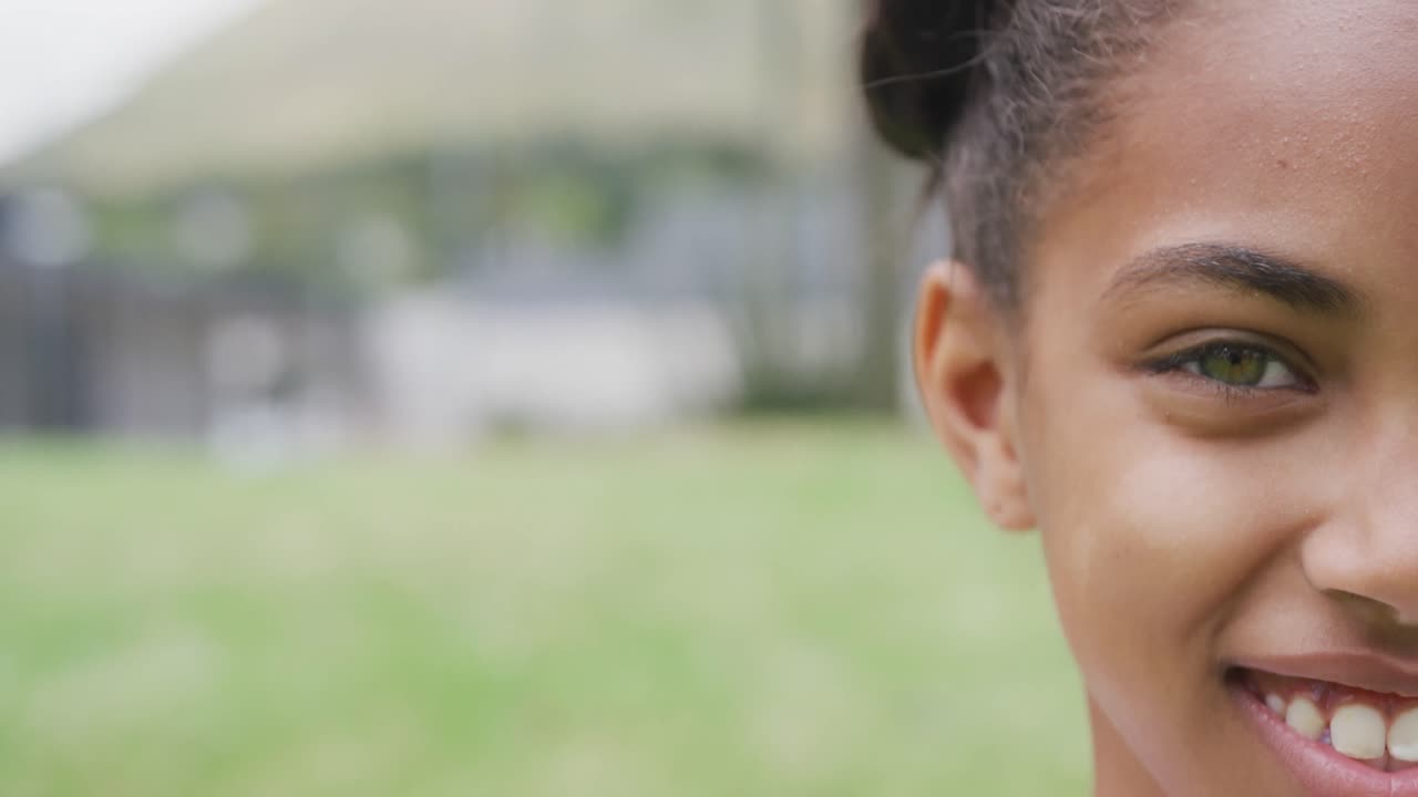 Video half face portrait of smiling biracial schoolgirl in school playing field, with copy space