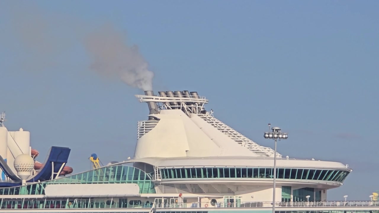 Närbild av övre däck och skorsten på ett stort lyxkryssningsfartyg som släpper ut grå avgasrök mot en klarblå himmel vid Port Everglades, Florida