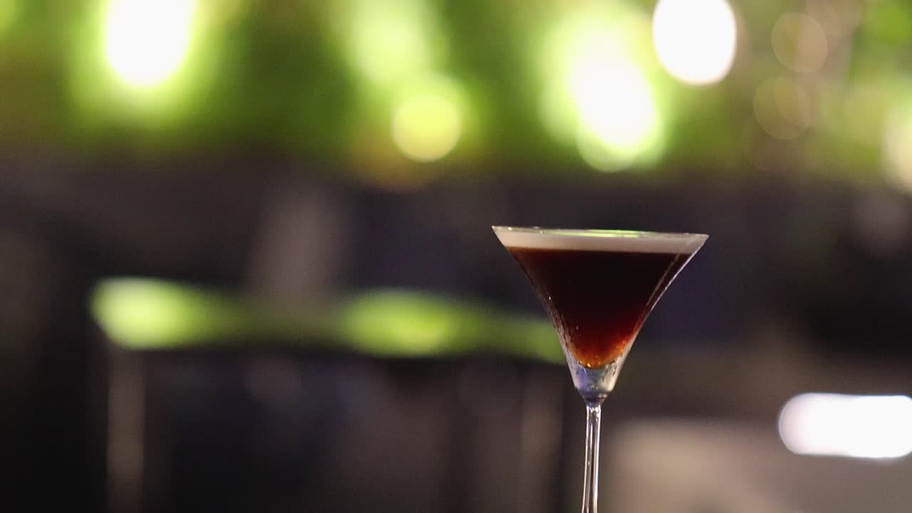 A person approaches an espresso martini on a table in a softly lit outdoor lounge