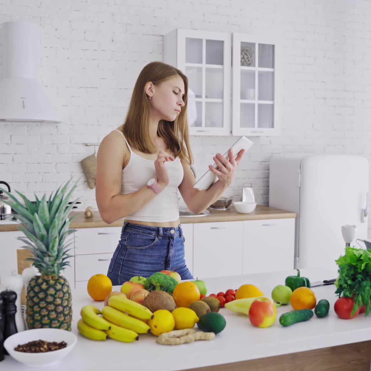 Pretty woman holds tablet in the kitchen. Young female is thinking about a new recipe of organic food. Fresh fruit and vegetables on the table. Healthy eating.