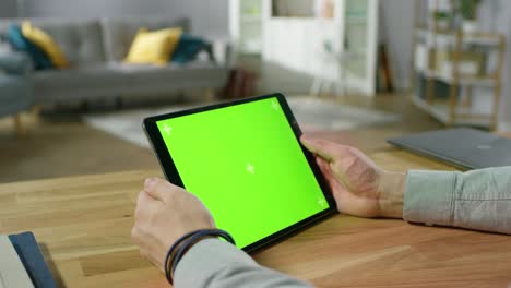 man holding and watching green mock-up screen digital tablet computer while sitting at the desk. man watching videos or browsing through the internet. in the background cozy living room.