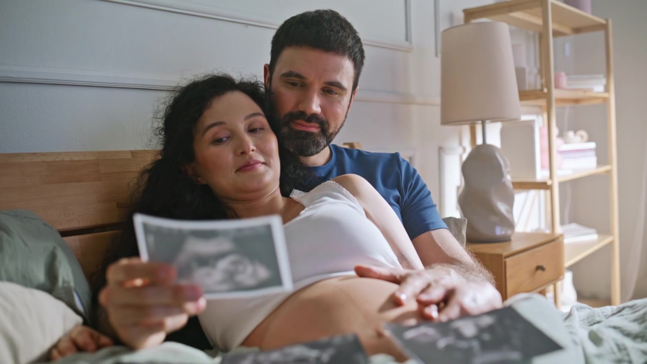 Expectant family viewing baby sonogram bed. Man embracing pregnant wife bedroom.