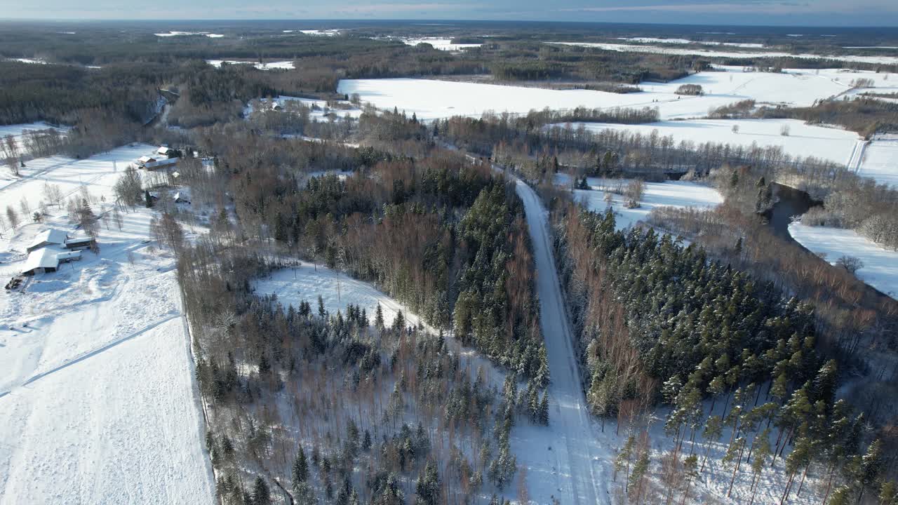Aerial view of a serene winter countryside featuring snow-covered trees, wooden houses, and a winding road through a frosty forest.