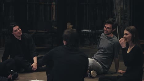 Close-up-a-group-of-confident-young-actors-in-black-suits-together-with-their-stage-director-in-a-gray-T-shirt-sit-in-a-circle-on-the-stage-and-communicate-with-each-other-during-a-rehearsal-for-a-performance-in-the-theater