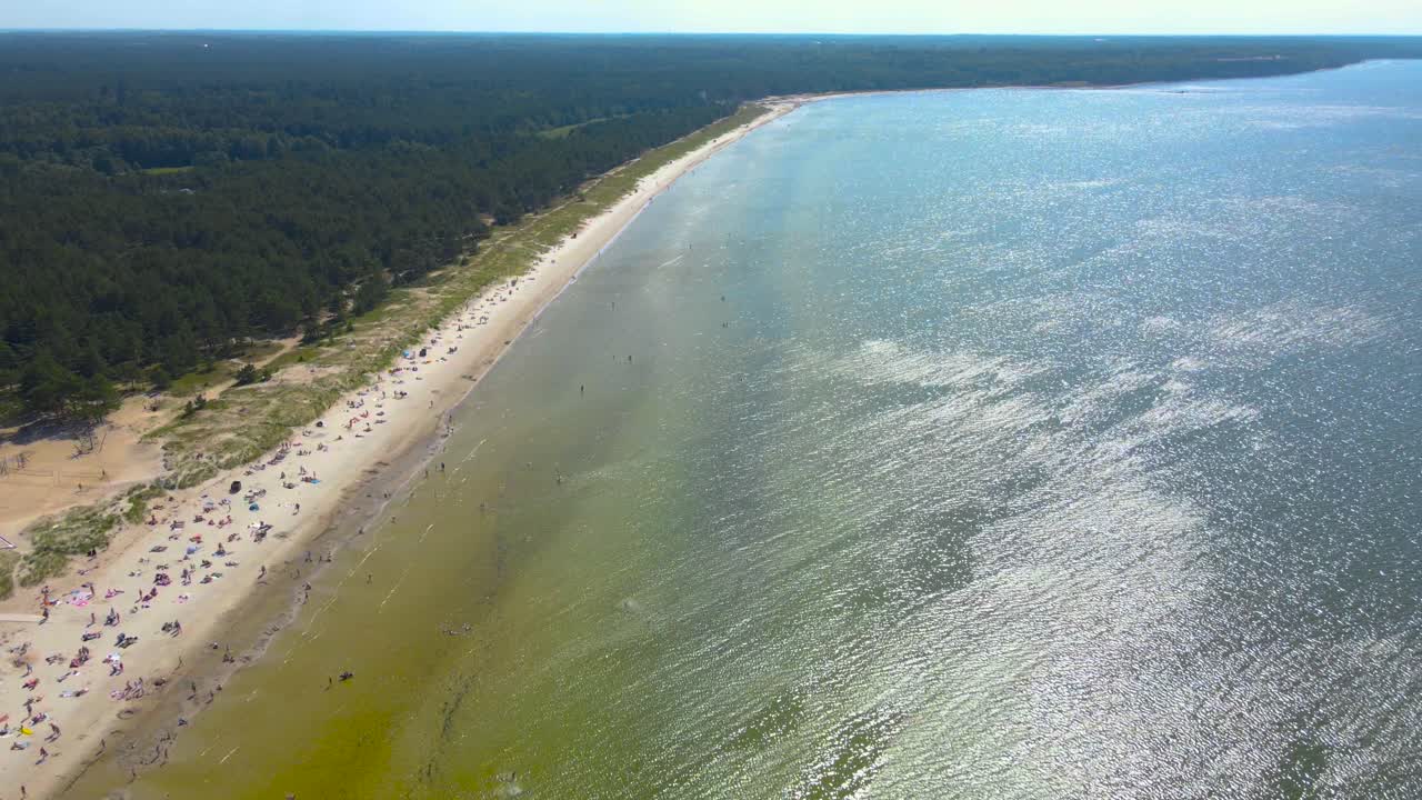 High altitude aerial drone footage flying over a sandy and people filled ocean sea shoreline beach during a summer hot and sunny day when people are sunbathing and swimming in the wavy blue water