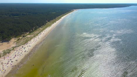 high altitude aerial drone footage flying over a sandy and people filled ocean sea shoreline beach during a summer hot and sunny day when people are sunbathing and swimming in the wavy blue water