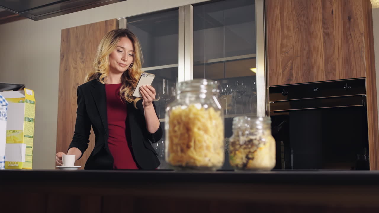 Una donna caucasica sorridente con i capelli biondi e una giacca beve caffè e guarda il telefono in cucina. Tavolo con cibo
