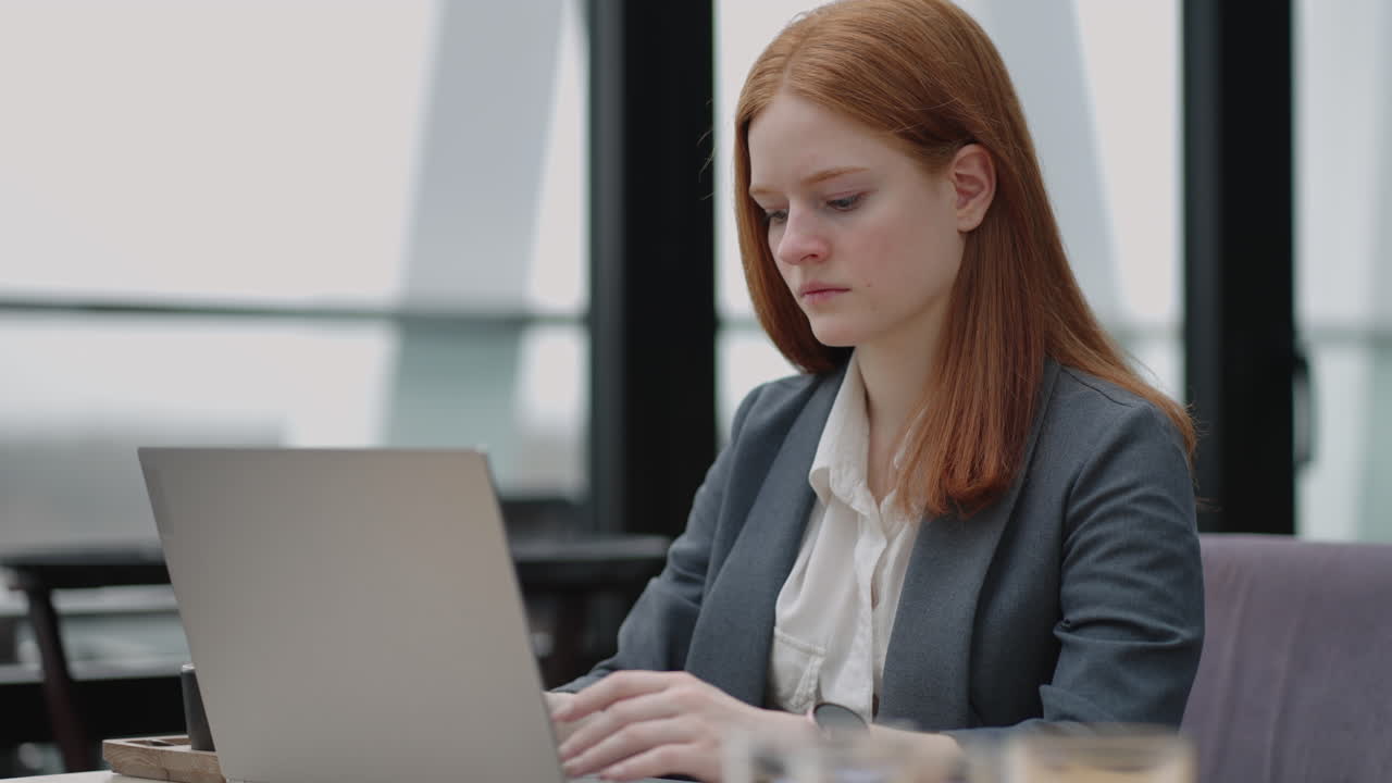 A young red-haired woman is working on a laptop. Remote work. Home office. Office work. A woman in a suit in the office is typing on a laptop