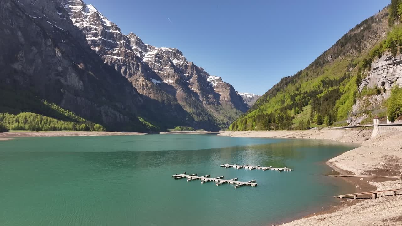 Aerial view of Klöntalersee in Glarus, Switzerland, with turquoise alpine waters, wooden docks, and majestic snow-capped mountains under a clear blue sky