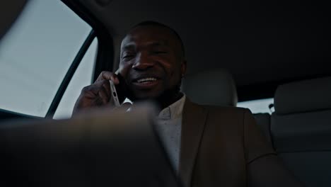 happy man businessman with black skin color in a brown suit reviews his plans on a tablet and reports them to his colleague using a phone while driving in a modern car outside an urban area