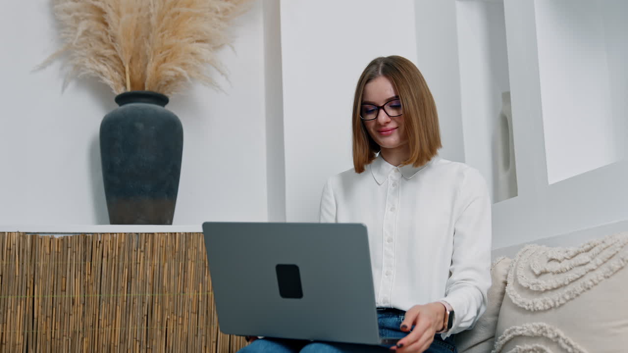 Comfortable workplace at home. Brunette short-haired woman wearing glasses sits working on her laptop. Low angle view.
