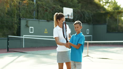 Mujer-Sonriente-Enseñando-A-Su-Hijo-Cómo-Golpear-La-Pelota-Con-La-Raqueta-En-Una-Cancha-De-Tenis