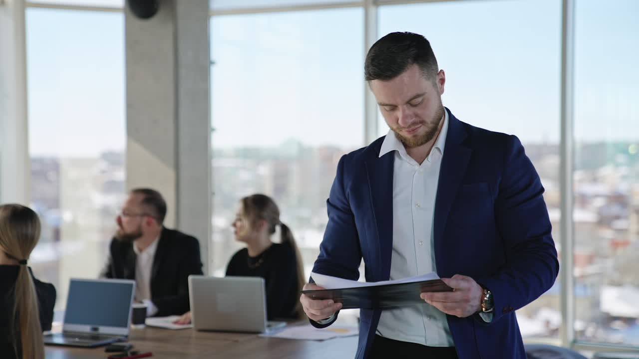 Young businessman in a suit holding the papers and looking at them. Young entrepreneur among the business partners at work. Team of coworkers at the background.
