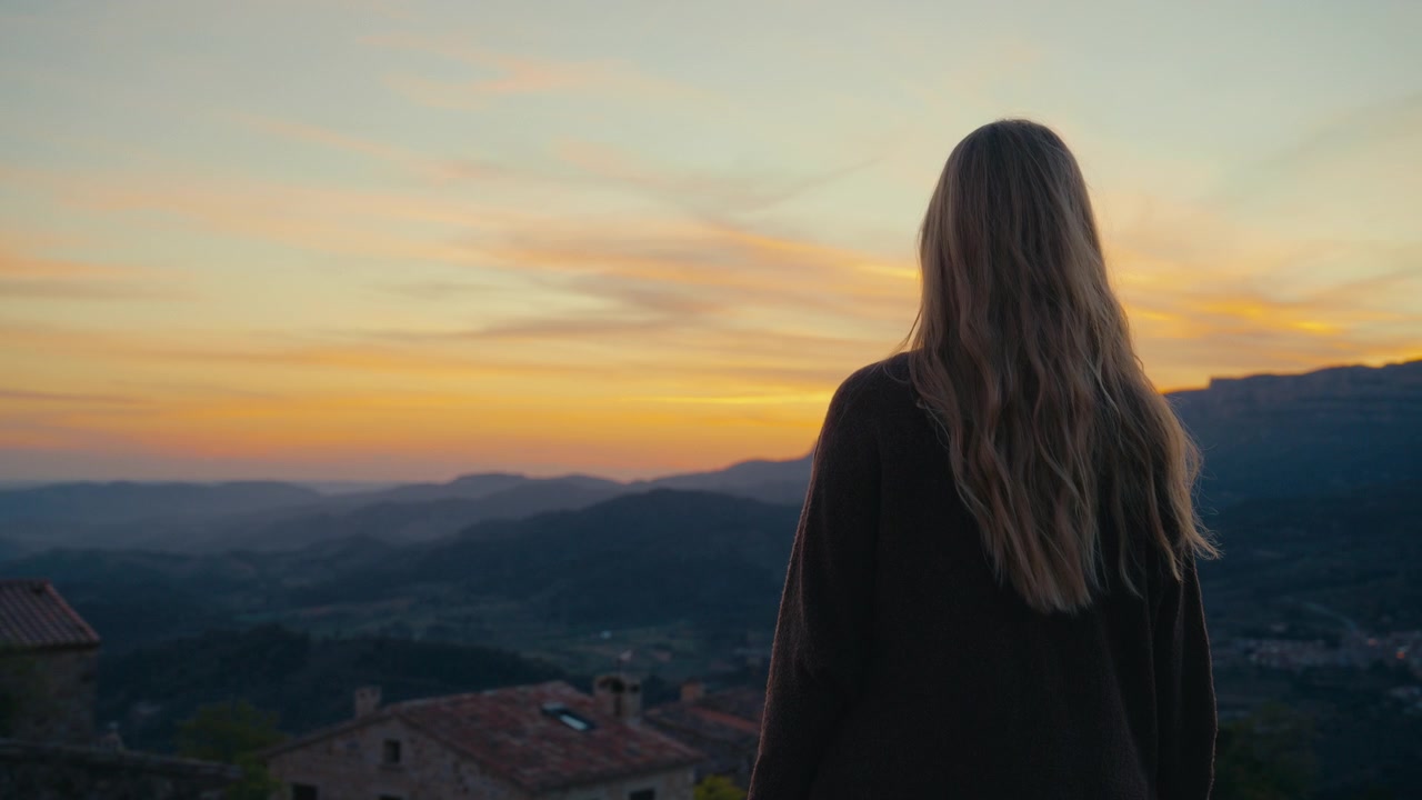 Una donna sta in piedi in silenzio sopra Siurana, Spagna, mentre i tenui colori del tramonto inondano il cielo e le colline lontane, creando una scena serale serena piena di luce calda e un'atmosfera calma.