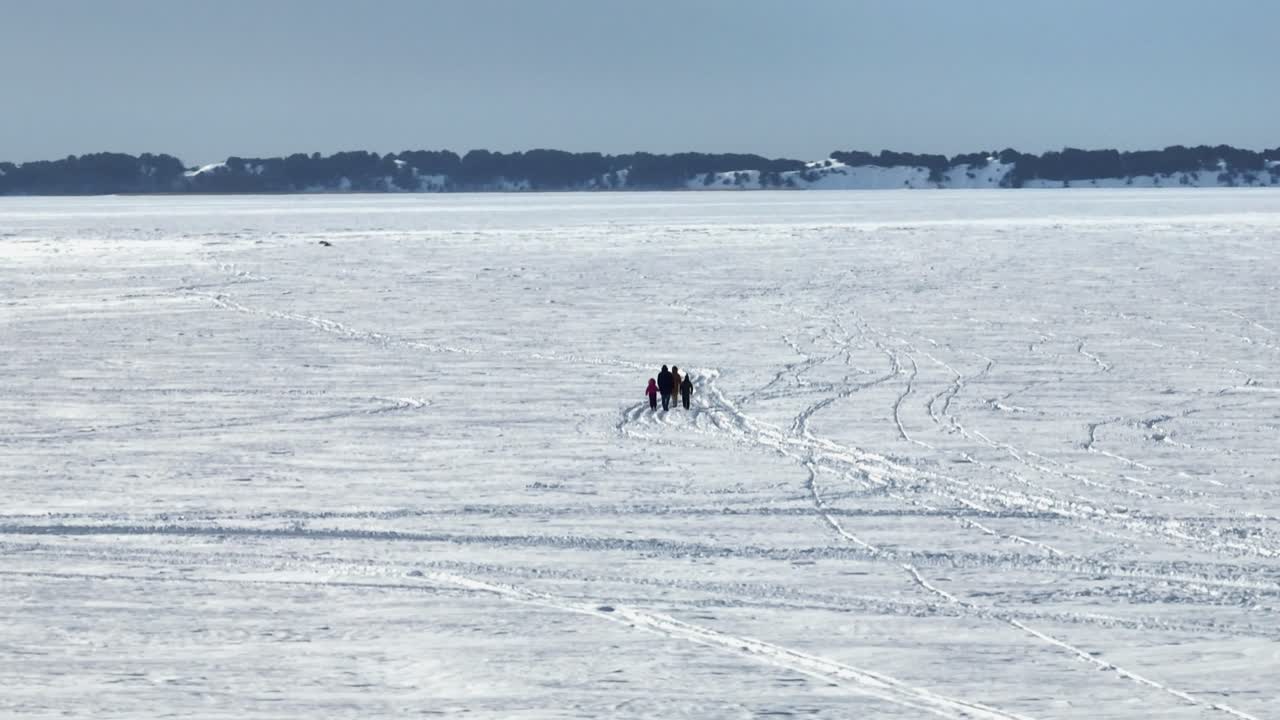 Uma vista distante, ao nível do solo, de um pequeno grupo a fazer caminhada através de um lago congelado e maciço, polvilhado de neve. Inúmeras pegadas no gelo levam em direção a um horizonte escuro e florestado sob um céu invernal nebuloso.