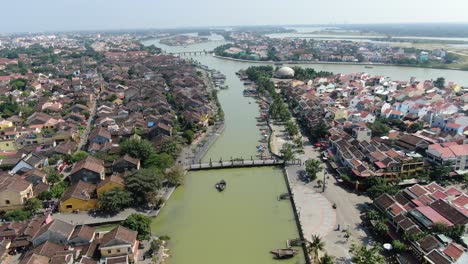 Drone-aerial-view-in-Vietnam-flying-over-Hoi-An-brown-color-river-canal-in-the-city,-small-brick-houses-and-wooden-boats-on-a-sunny-day