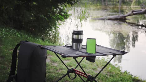 Camping table with thermo cup and smartphone outdoors