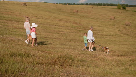 woman and children walking down a grassy hill with a dog on a leash, the young girl carries a basket while the boy follows closely, the scenic background features rolling fields and hay