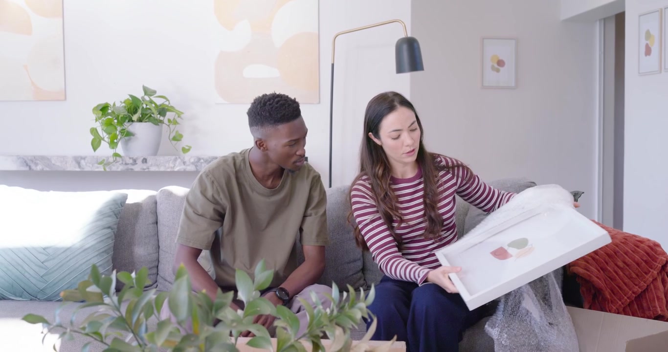 Casal diverso desempacotando caixas de papelão, desembrulhando plástico bolha, inspecionando uma impressão e decorando a casa.