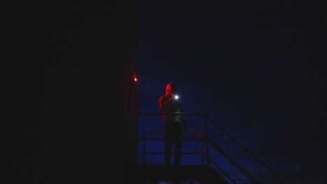 mysterious hooded man on top of metal stairs shining a flashlight around at night time