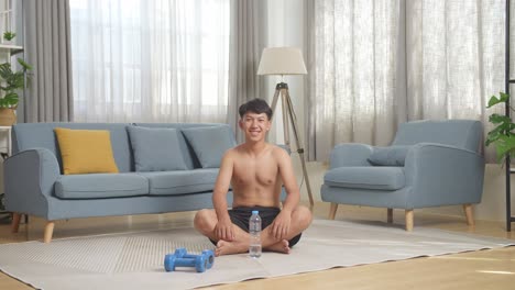full body of asian teenager boy with dumbbells smiling to camera while sitting on mat during the shirtless workout at home