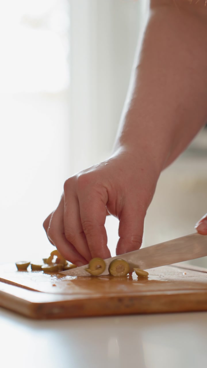 Una vista dal basso mostra una mano adulta che trita piccoli ingredienti rotondi su un caldo tagliere di legno, con la lama del coltello che preme mentre le punte delle dita guidano i pezzi in una luminosa cucina di casa, con mano e coltello.