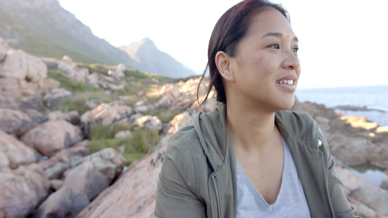 Smiling woman enjoying mountain hike, sitting on rocky terrain near ocean