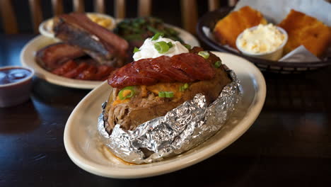 Loaded-baked-potato-with-smoked-sausage,-traditional-barbecue-plate-and-cornbread-in-background,-slider-close-up-4K