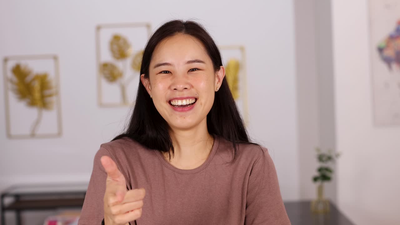 Young Asian woman smiles and points at the camera with a cheerful expression in a well-lit, modern indoor setting with soft background decor