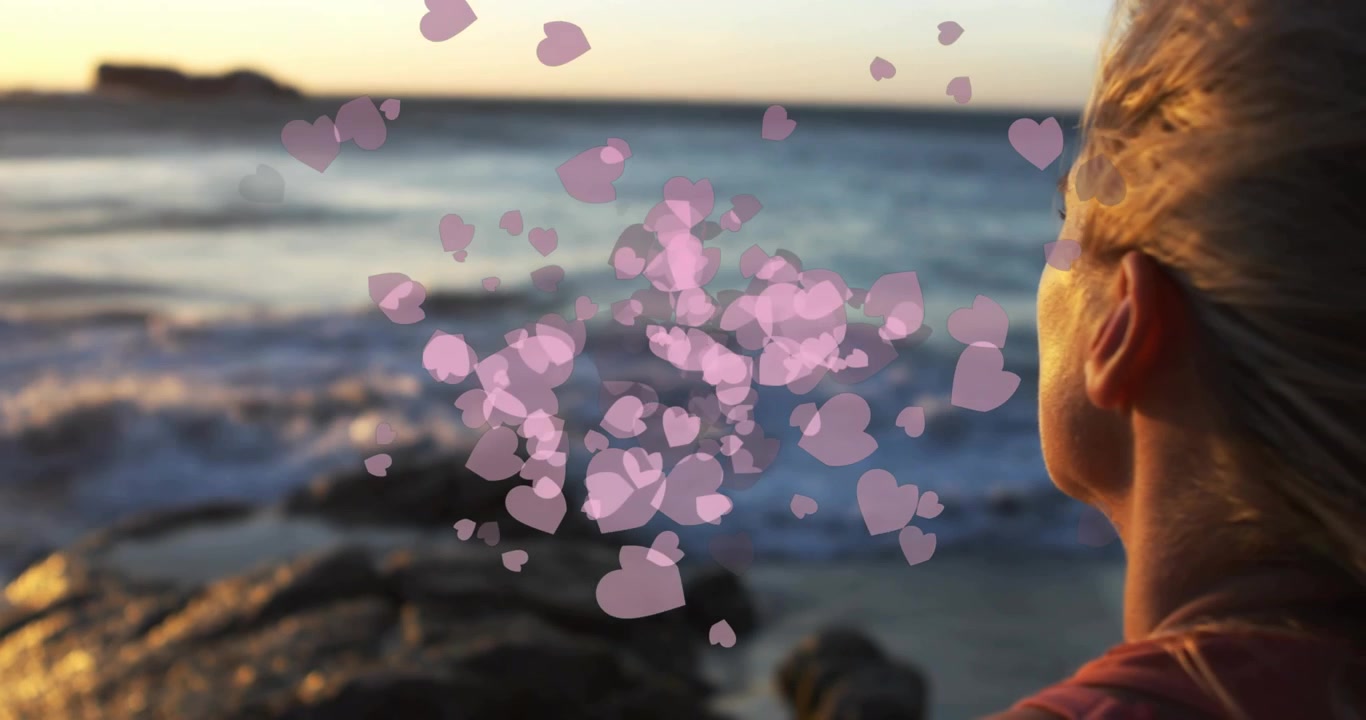 Sonnenuntergangsbeleuchtung mit rosa Herzen, die sich um eine Frau, die aufs Meer blickt, gruppieren und zerstreuen – für Marketingzwecke.