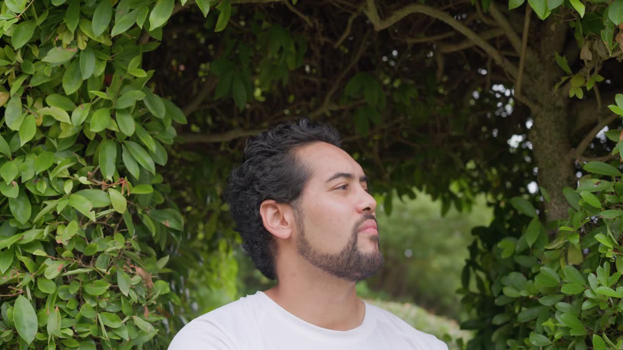 Uomo afroamericano in piedi in un giardino con arco di siepi, ripreso dalla telecamera, che sposta lo sguardo e sorride, indossando una maglietta bianca.