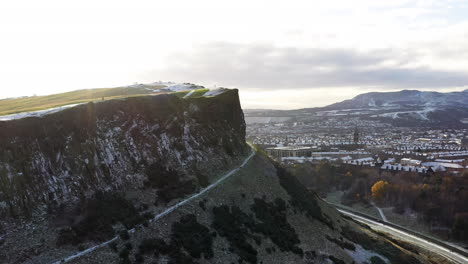 Aerial-shot-flying-around-Salisbury-crags-in-Edinburgh,-revealing-the-city-below-covered-in-snow-on-a-sunny-day