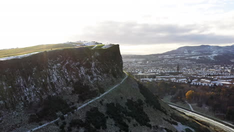 Toma-Aérea-Volando-Alrededor-De-Los-Riscos-De-Salisbury-En-Edimburgo,-Revelando-La-Ciudad-Debajo-Cubierta-De-Nieve-En-Un-Día-Soleado