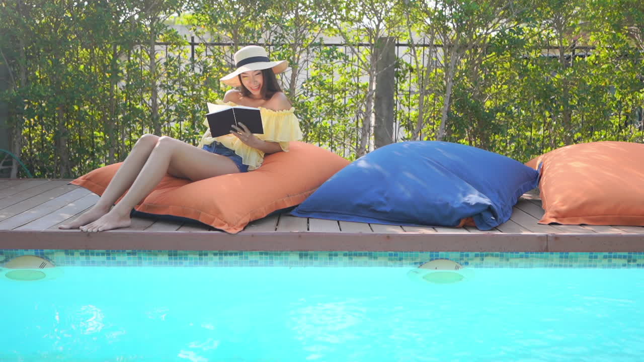 A pretty young woman sits poolside on a beanbag while reading a book
