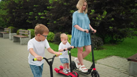 a mother and her two children ride scooters outdoors, the younger child glances at his older brother as they ride side by side