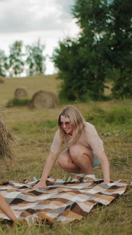 dama y niño extienden la tela para un picnic cerca de la bala de heno con un libro colocado en la parte superior, vista parcial de otra persona y perro al lado en una vasta tierra de cultivo