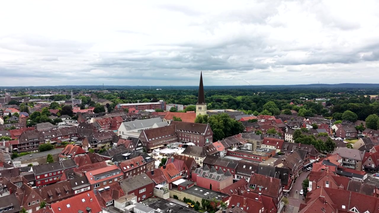 Ein hoher dunkler Kirchturm durchstößt den bedeckten, bewölkten Himmel über dichten rotgedeckten Dächern in Dülmen, Deutschland. Schmale, von Bäumen gesäumte Straßen schlängeln sich durch das historische Altstadtzentrum. Weitwinkelaufnahme mit Drohne.