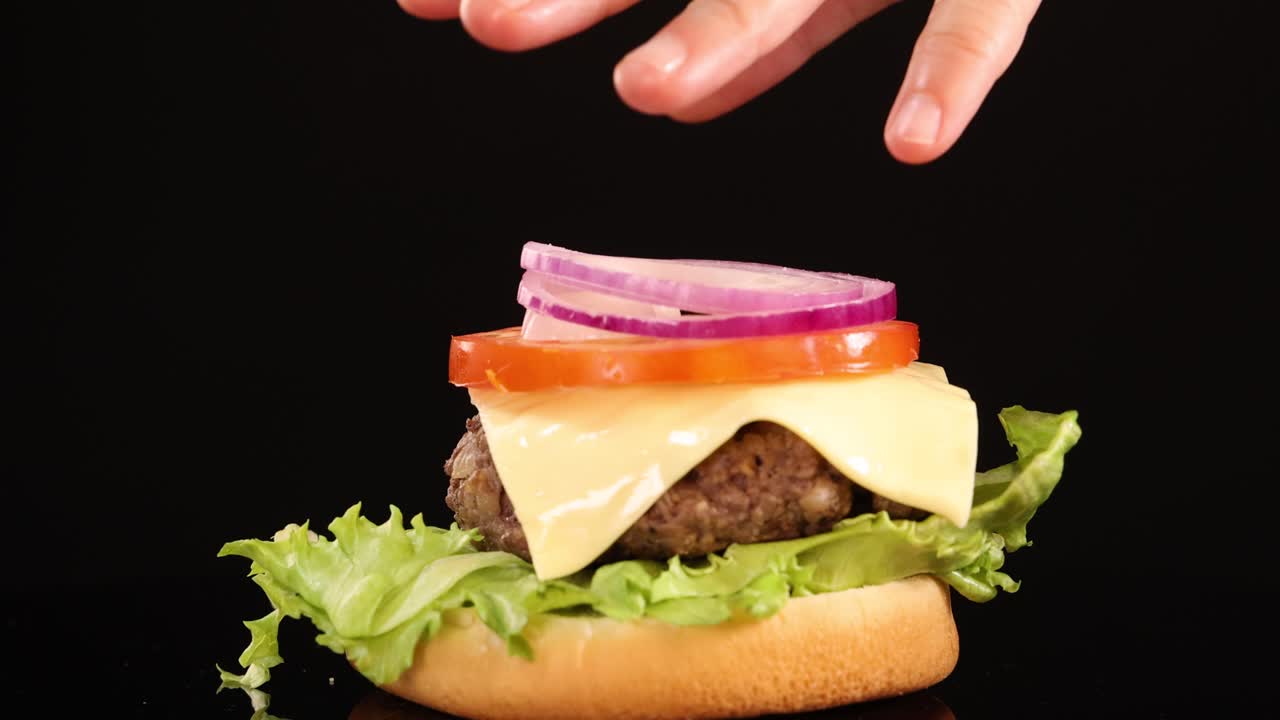 Hands layer tomato, onion, cheese, and lettuce onto a beef patty and bun under bright studio lighting, finishing with a sesame seed top bun